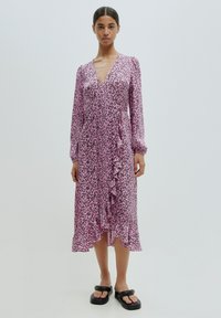 Rosa Blumenwickelkleid mit langen Ärmeln, gerafftem Vorderteil und einem mittellangen Saum, aus leichtem Stoff; kombiniert mit schwarzen Plateausandalen.