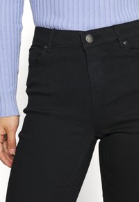 Jeans negros de cintura alta con una tela suave y elástica. Presentan un cierre de botón metálico y bolsillos frontales, combinados con una blusa de canalé azul claro.