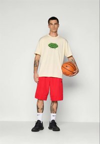 Krém színű, oversize póló zöld Nike logóval, piros sportnadrág, fekete sportcipő, kosárlabdát tartva. Nike logós rövid zokni.