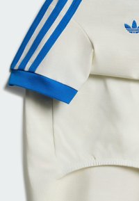 Tissu blanc avec poignet bleu et trois bandes bleues sur la manche, avec un logo partiel Adidas bleu près de l'épaule.