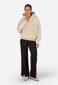 Beżowy hoodie z kieszenią z przodu i sznurkami, w połączeniu z czarnymi spodniami typu wide-leg z bocznymi rozporkami, noszony z beżowymi sneakerami.