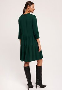 Zielona sukienka z rękawami o długości trzech czwartek, warstwowym dołem i gładkiego materiału. W zestawieniu z czarnymi, za kolano stiletto, z ostro zakończonymi noskami.