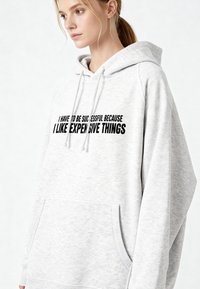 Felpa grigia con cappuccio dotato di coulisse, tasca a marsupio e testo nero: "DEVO AVERE SUCCESSO PERCHÉ MI PIACCIONO LE COSE COSTOSE."