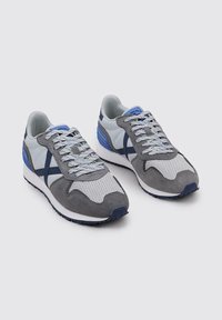 Zapatillas deportivas grises y blancas con materiales de malla y ante, acentos azules, lengüeta texturizada y sistema de cordones. Suela de goma con relieve.