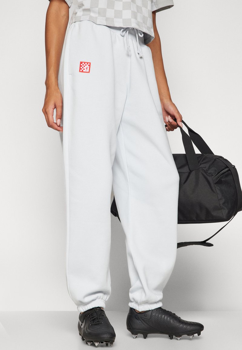 Pantaloni da tuta grigi chiari con polsini elastici, caratterizzati da un logo a scacchi rosso e bianco, abbinati a scarpe da calcio nere e a una borsa a tracolla nera.