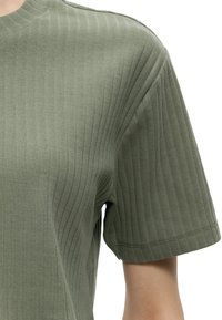 Close-up of a green ribbed short-sleeve top on a person’s upper torso and arm against a white background.