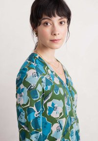 Blusa verde con estampado floral que presenta grandes flores azules y blancas, escote en V con detalle de botones y textura de tela suave.