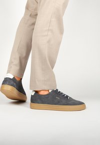 Zapatillas de ante gris con suela de goma, cordones en color marino y detalles en blanco en el talón, combinadas con unos pantalones beige claros de corte ajustado.