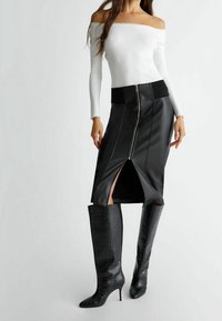Jupe midi en cuir noir avec fermeture éclair à l'avant, fentes latérales et large taille noire, associée à des bottes noires texturées montantes jusqu'aux genoux. Haut blanc épaules dénudées.