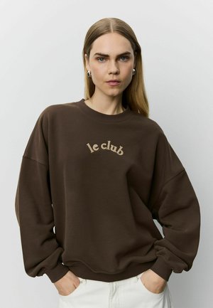 Donna con capelli biondi lisci che indossa orecchini a cerchio argentati, una felpa marrone con testo "le club" e pantaloni bianchi, mani in tasca.