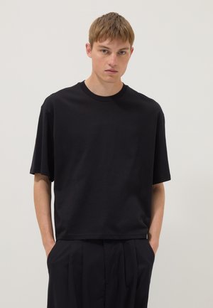 Jonge man met kort haar die een effen zwarte oversized T-shirt en zwarte broek draagt, staande tegen een lichte achtergrond.