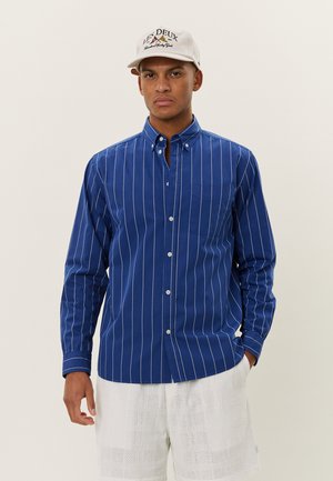 Blaues Hemd mit Knopfleiste und weißen vertikalen Streifen, strukturierter Stoff und einer Brusttasche. Getragen mit hellen, gemusterten Shorts und einer beigen Mütze.