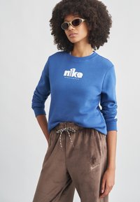 Μπλε μπλούζα φούτερ με λαιμόκοψη crewneck και λευκό λογότυπο Nike, σε συνδυασμό με καφέ, μαλακές φόρμες με κορδόνι στη μέση και χρυσή λεπτομέρεια λογότυπου.