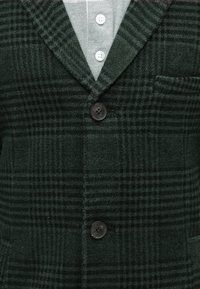 Tmavě zelený kostkovaný blazer s výstřihem do V, jedním knoflíkovým zapínáním a kapsou. Měkká, strukturovaná tkanina s kostkovaným vzorem.