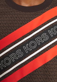 Gros plan sur un tissu marron avec un motif répétitif de petits "MK", présentant des rayures diagonales rouges et noires avec le texte "KORS" en contours blancs gras.