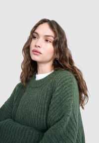 Jeune femme aux cheveux bruns ondulés portant un pull vert texturé par-dessus un haut blanc, regardant pensivement sur le côté sur un fond gris clair.