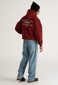 Mand iført en bordeaux hættetrøje med hvid tekst på ryggen, lyseblå løse jeans, sorte sko og en sort strikket beanie, står oprejst foran en ensfarvet baggrund.