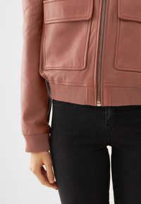Giacca bomber in pelle rosa con tasche frontali, polsini a costine e zip completa. Abbinata a jeans neri, dal design elegante e minimal.