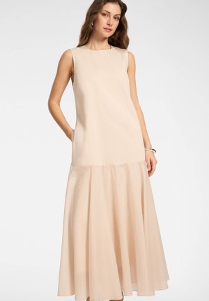 Vrouw draagt een mouwloze, beige, vloerlang jurk met uitlopende zoom, staand met één hand in de zak tegen een effen achtergrond.