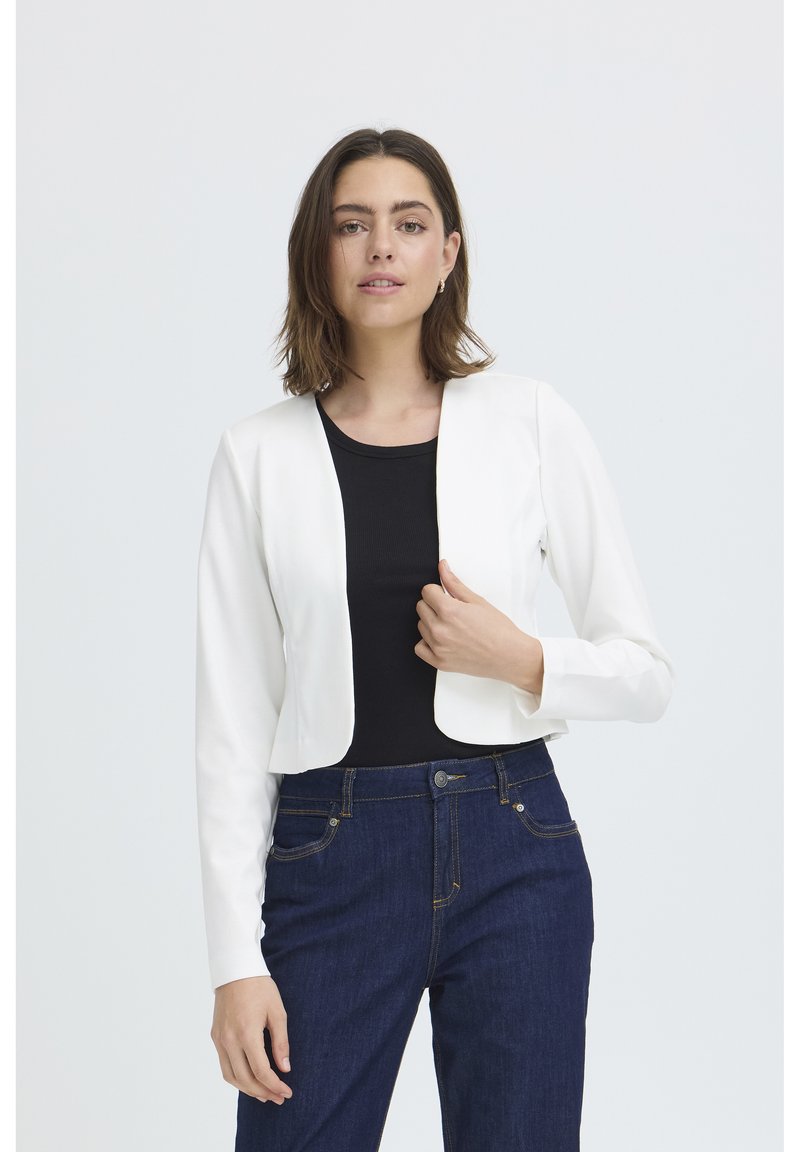 Femme aux cheveux bruns mi-longs porte un blazer blanc court sur un haut noir et un jean bleu foncé, debout devant un fond uni.
