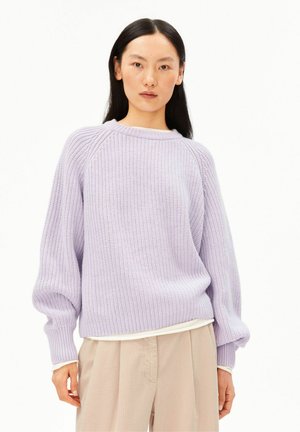 Femme aux longs cheveux noirs portant un pull en tricot violet clair et un pantalon beige, debout devant un fond blanc uni.