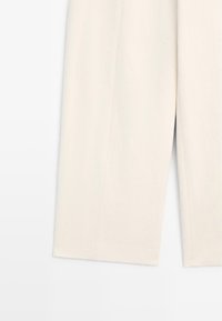 Pantalon beige clair à jambes larges avec une fente à l'avant, montré de la mi-cuisse à l'ourlet sur un fond blanc uni.