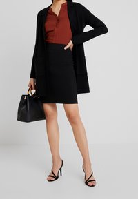 Giacca nera con tasche, blusa color ruggine, gonna nera aderente, tacchi aperti neri e una borsa nera con manici in legno.