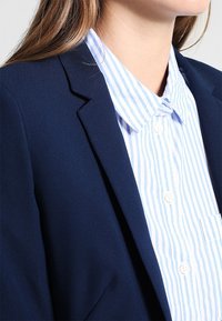 Mujer con un blazer azul sobre una camisa blanca con rayas verticales de color azul claro, mostrada de los hombros a la barbilla.