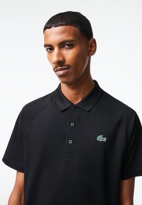 Černé polo tričko s tříknoflíkovou légou, krátkými raglánovými rukávy a zeleným vyšitým logem krokodýla. Hladká, texturovaná látka.