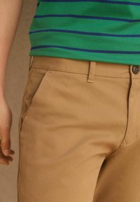 Gros plan d'une personne portant un pantalon beige et une chemise verte avec des rayures horizontales bleues, mettant l'accent sur la taille et la zone de la poche.
