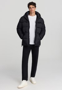 Giacca a vento nera con cappuccio, caratterizzata da un design trapuntato e tasche frontali. Indossata sopra una camicia bianca, abbinata a pantaloni neri e scarpe bianche.