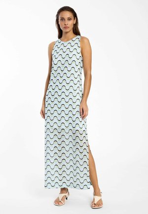 Vrouw staand, gekleed in een mouwloze maxi-jurk met blauw-zwarte golvende strepen en beige open-teen sandalen met hak, tegen een witte achtergrond.