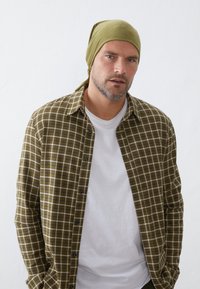 Grønternet skjorte med sorte knapper over en hvid t-shirt, parret med et matchende grønt hovedklæde, sat op mod en ensfarvet baggrund.