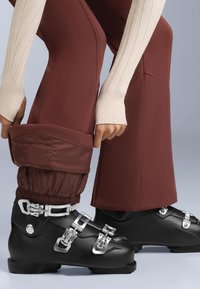 Čierne lyžiarske topánky so striebornými prackami, v kombinácii s gaštanovými nohavicami s riasenými manžetami a textúrovaným krémovým sveterom. Zamerajte sa na detaily.