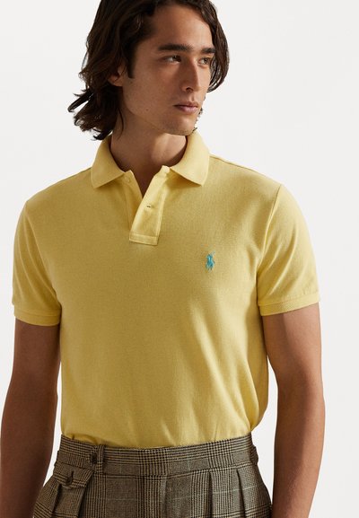 Dzeltena polo krekla no tekstūrētas kokvilnas ar trīs pogu apiešanu un tirkīza krāsas logo kreisajā krūtī. Savienots ar rakstainām biksēm.