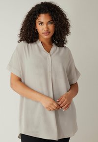 Blusa gris clara con un sutil patrón de cuadrícula, mangas cortas, escote en V y cierre de tres botones en el frente. Textura suave.