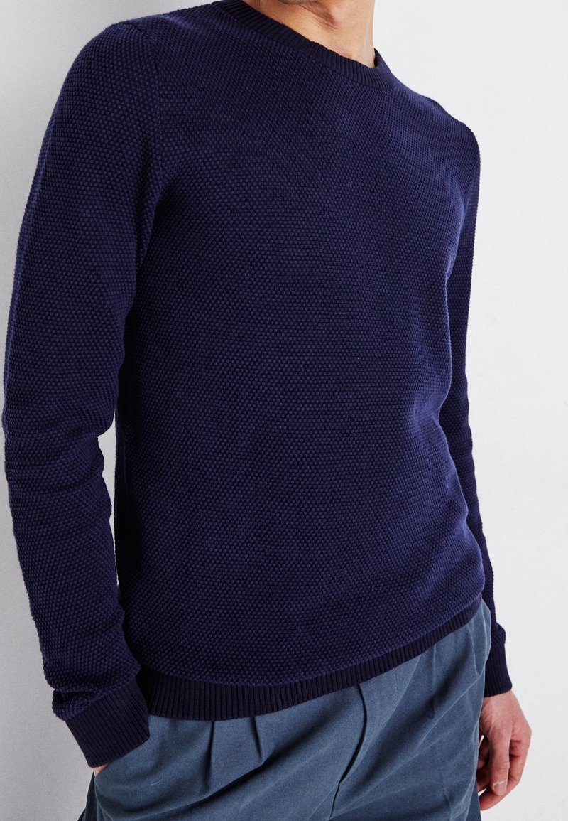 Marinblå stickad tröja med texturerad tyg, rund halsringning och ribbad nederkant och ärmslut. Matchas med ljusblå byxor.