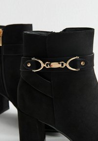 Botas de tobillo de ante negro con un detalle de hebilla y correa de metal dorado. Tacones anchos y resistentes con forro interior suave.
