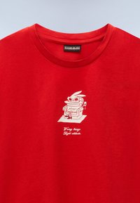 Κόκκινο μπλουζάκι με ένα μικρό λευκό γραφικό πουλιού με γυαλιά ηλίου, καθισμένο σε πολυθρόνα και κρατώντας ένα ποτήρι, με κείμενο από κάτω.