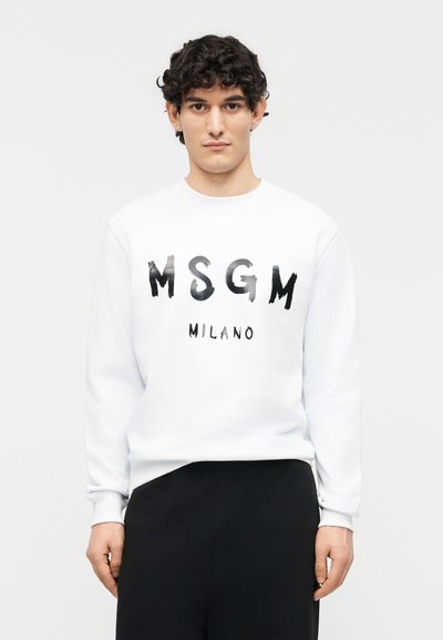 Jeune homme aux cheveux bouclés portant un sweat-shirt blanc MSGM Milano et un pantalon noir, debout devant un fond blanc uni.