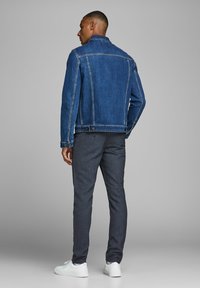 Giacca di jeans blu scuro con taglio classico, dotata di tasche frontali con bottoni e cuciture a contrasto. Abbinata a pantaloni grigi con trama e sneakers bianche.
