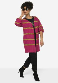 Gebreide cardigan in helder roze met gele en groene strepen, met een open voorkant, lange mouwen en geribde boorden, gedragen over een zwarte top en broek.