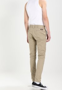 Hnědé, slim-fit kalhoty vyrobené z hladké látky, s kapsami na zadní straně a malou nášivkou, kombinované s bílým tílkem a černými teniskami.