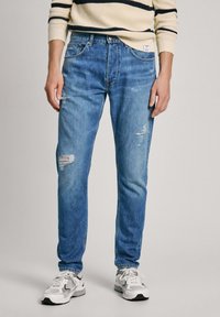 Vaaleansiniset slim fit -farkut, joissa on näkyviä repeämiä, haalistuneita kohtia ja klassinen viiden taskun muotoilu. Käytetty valkoisten tennareiden kanssa.