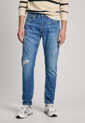 Gai zilganzs džinsi ar šauru piegriezumu, ar redzamām izgriezumiem, izbalējušām vietām un klasisku piecu kabatu dizainu. Valkāti ar baltiem sporta apaviem.