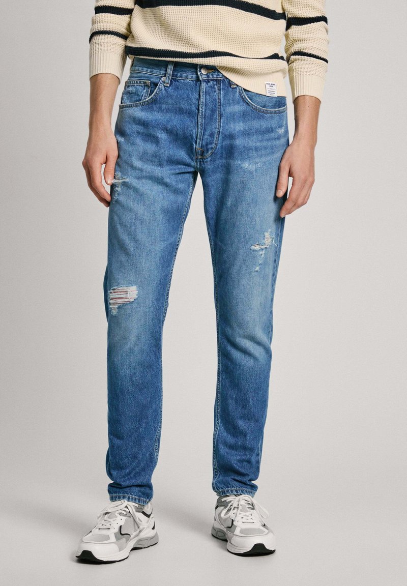 Vaaleansiniset slim fit -farkut, joissa on näkyviä repeämiä, haalistuneita kohtia ja klassinen viiden taskun muotoilu. Käytetty valkoisten tennareiden kanssa.