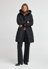 Svart vadderad lång kappa med huva, med dragkedjestängning, sidofickor och logotypdetaljer. Kombineras med svarta byxor och klackstövlar.