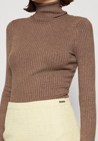 Bruine geribbelde coltrui met lange mouwen, gemaakt van een gestructureerde breistof. Gecombineerd met lichtgele geweven hoge taille broek.