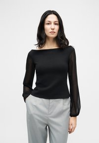 Γυναίκα με σκουρόχρωμα μαλλιά φοράει μαύρο τοπ έξω από τον ώμο με διάφανα μακριά μανίκια και ανοιχτό γκρι παντελόνι με ρίγες, όρθια μπροστά σε απλό φόντο.