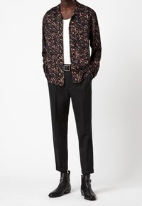 Camicia a maniche lunghe a fantasia nera e marrone indossata sopra una T-shirt bianca, abbinata a pantaloni neri su misura e stivaletti neri eleganti.